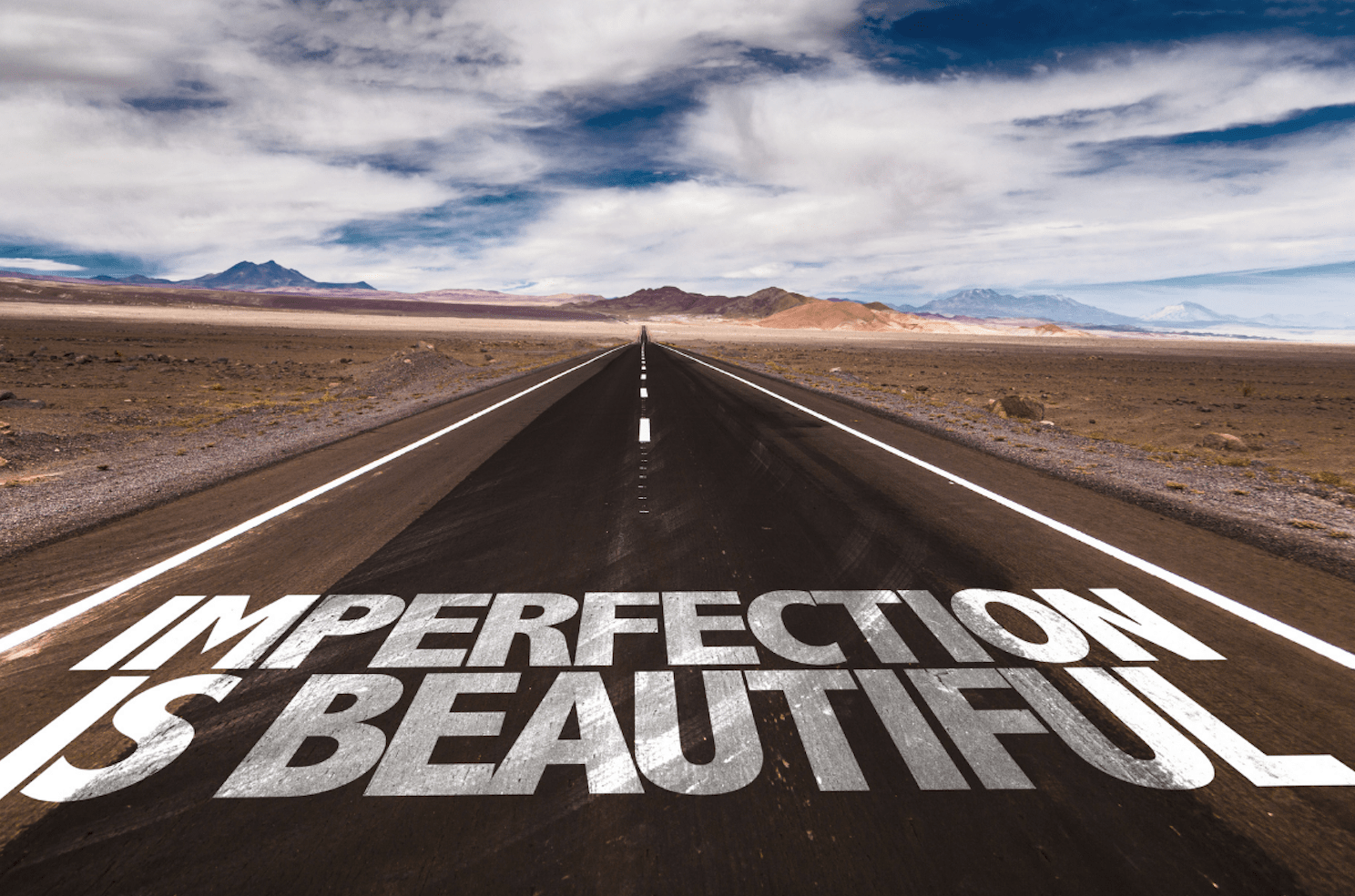 Imperfection is Beautiful painted on a road with mountains and cloudy day sky on the horizon.