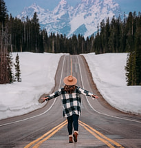 Woman walking down a road with no traffic and mountains that look like the Himalayas ahead. It is so gorgeous! Pine trees all around.