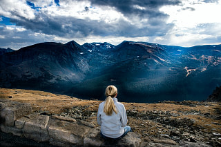 Lady meditating in a grassy area, overlooking a beautiful mountain ridge with the sun peering through a cloudy sky above.