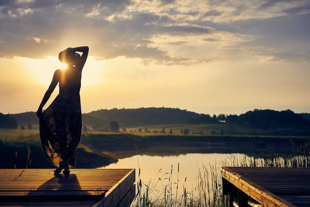 The back of a woman with a sunhat on, standing on a pier, looking over a lake with a beautiful sunset over a cascade of green hills.